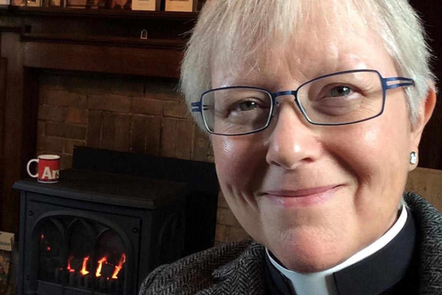 Cally is a white woman with white hair and glasses wearing a clerical collar under a black shirt and grey tweed jacket. She is in front of a mantlepiece decorated with card, framed photos and an Icon, and a lit log burner. She is smiling.