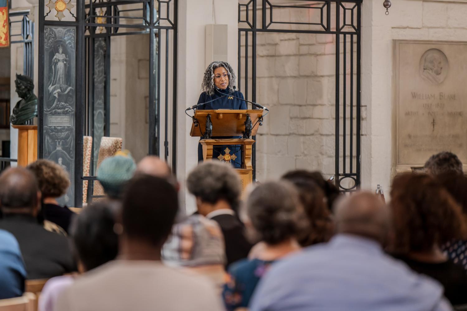 A black woman stands at a lectern speaking to a full audience