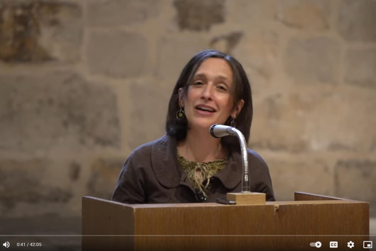 Susanna is a white woman with shoulder length dark hair standing at a microphone and lectern in front of an old stone wall inside the Cathedral