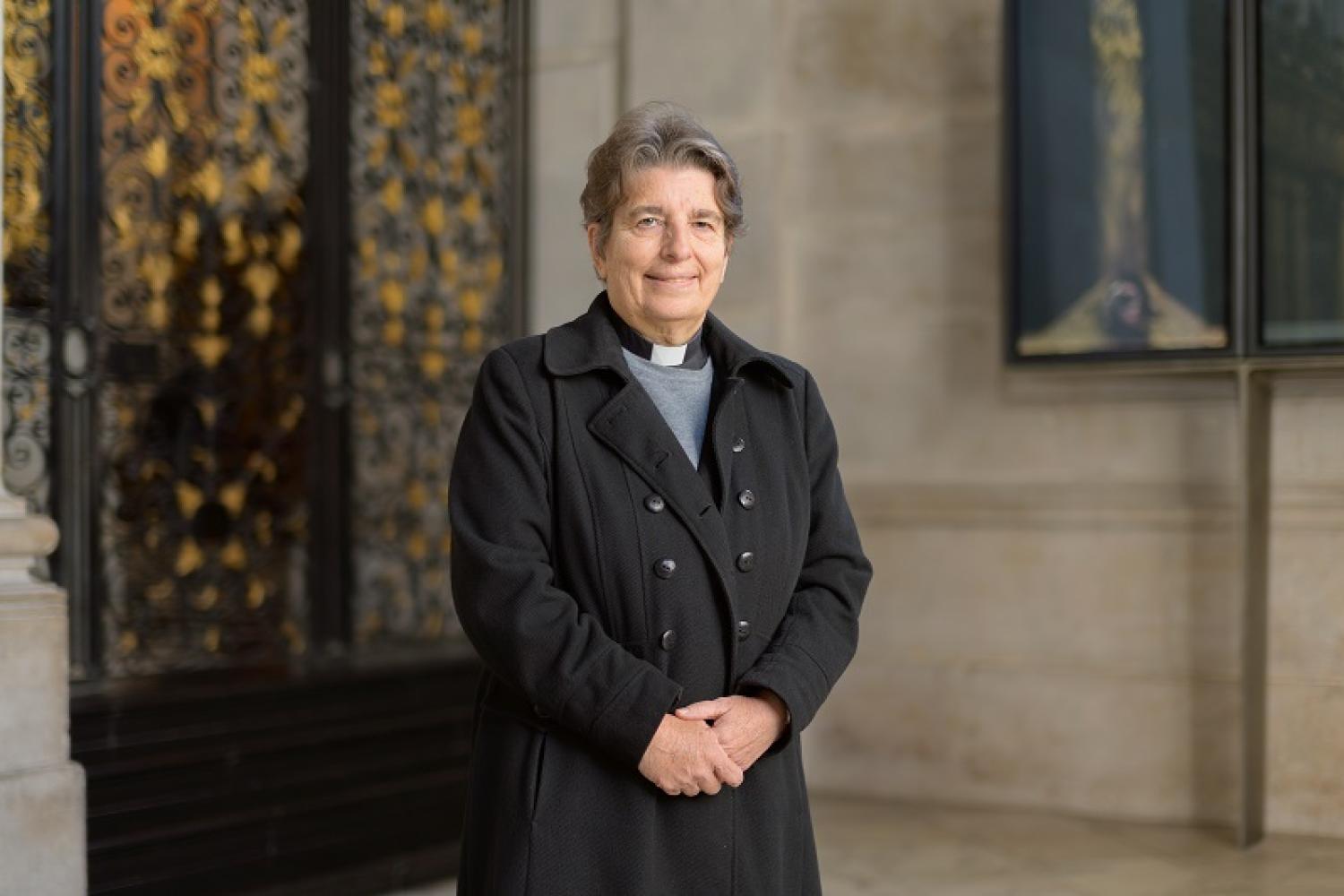 Paula Hollingsworth, a white woman with short dark hair in a clerical collar, stands in front of a wooden staircase.
