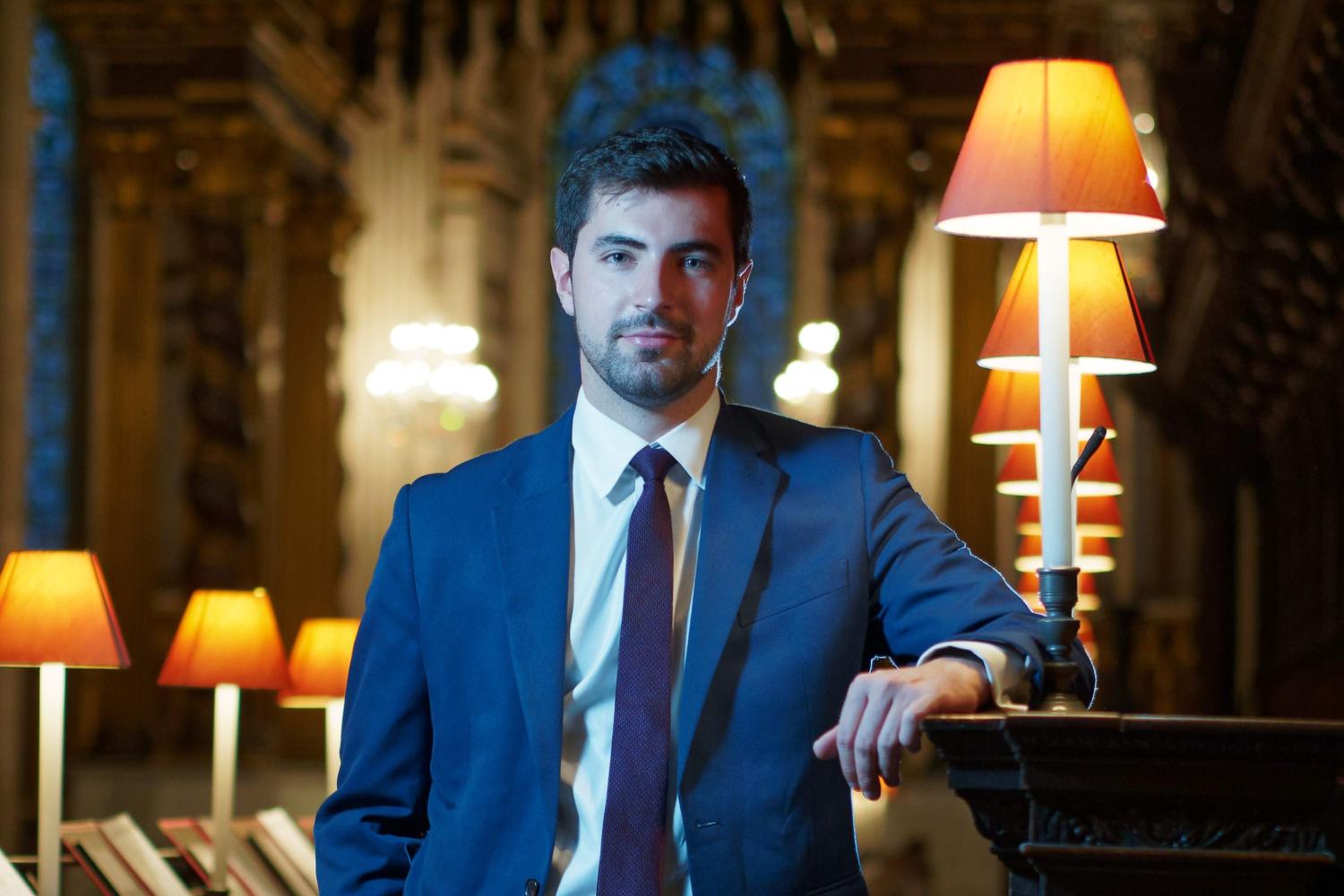 A portrait of James Orford. James is a white man with dark, short hair and a beard. He is wearing a suit and tie, and is stood in the Quire of St Paul's Cathedral.