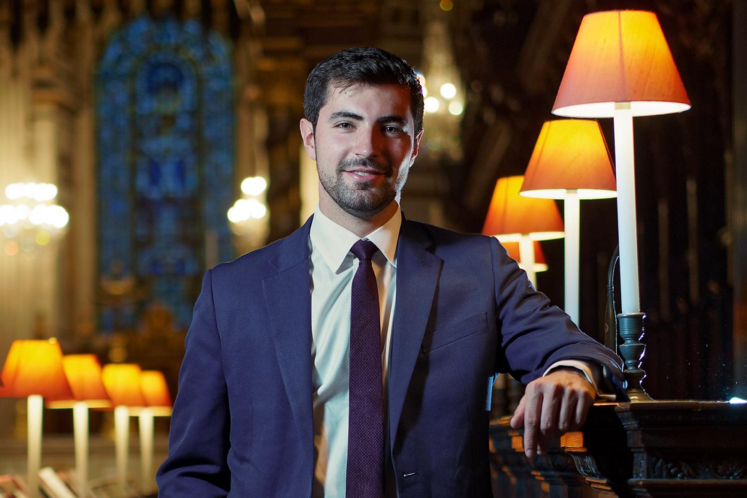 James is a white man with short dark hair wearing a blue suit. He stands in the quire at St Paul's Cathedral, leaning against a stall, with lit lamps alongside.