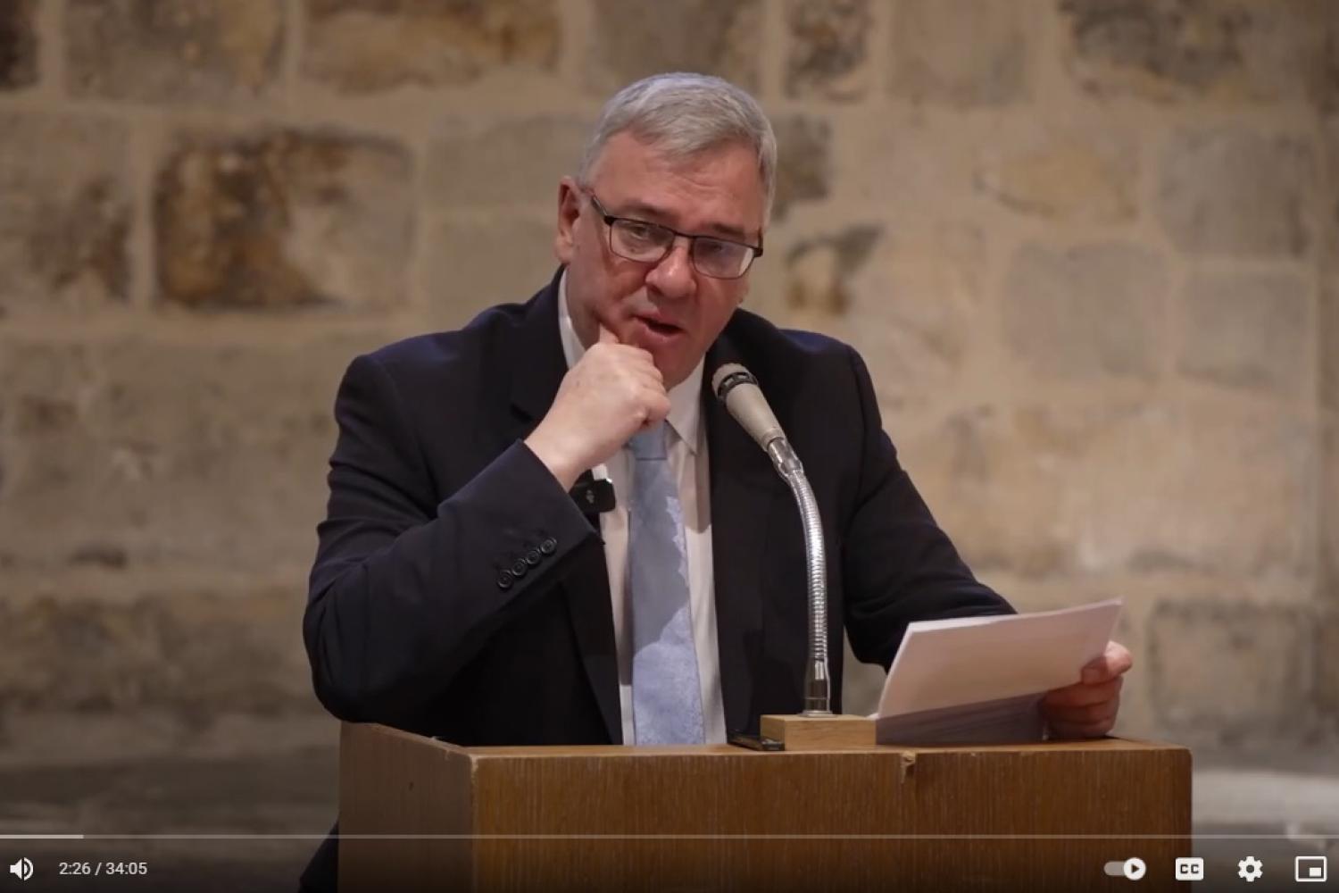 Andrew Carwood, a white man with grey hair and glasses, in a dark suite with a pale blue tie, stands at a microphone in front of a stone interior wall