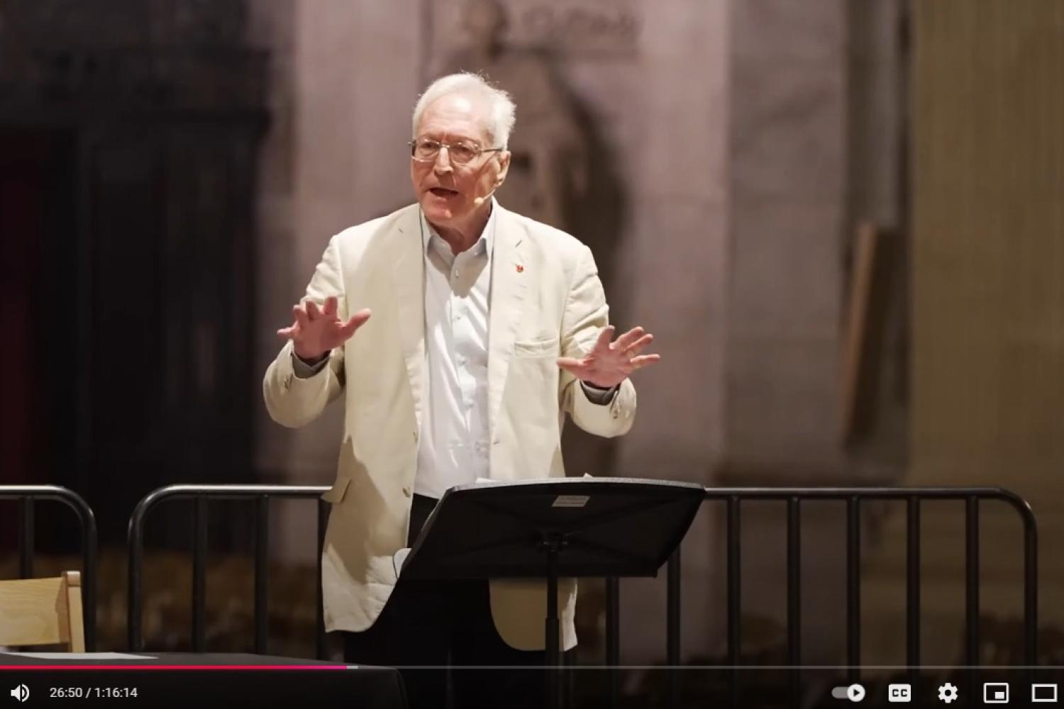 Diarmaid MacCulloch, a white man with white hair and glasses, wearing a cream linen jacket over a white shirt, stands at a lectern in St Paul's Cathedral