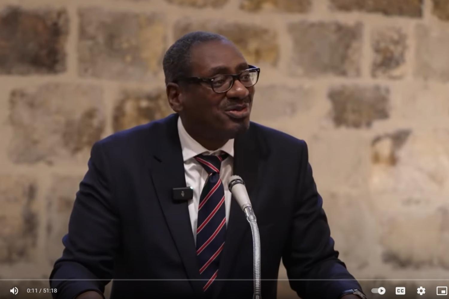 Richard is a black man with glasses wearing a navy suit and tie talking at a microphone in front of a stone wall