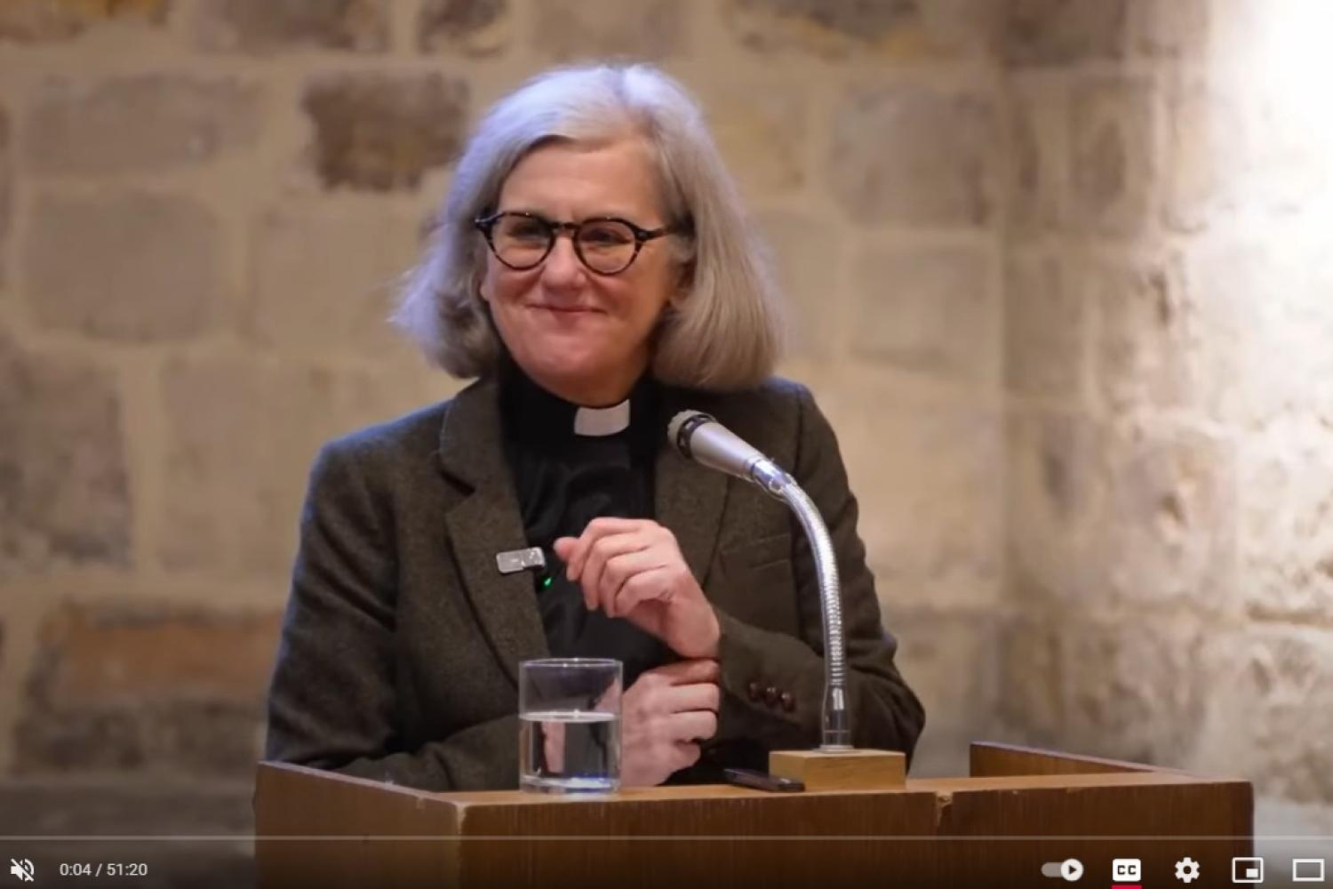 Janet is a white woman with grey bobbed hair and black rimmed glasses wearing a clerical collar and black shirt under a dark grey jacket, standing in front of a lectern and microphone and an interior stone wall