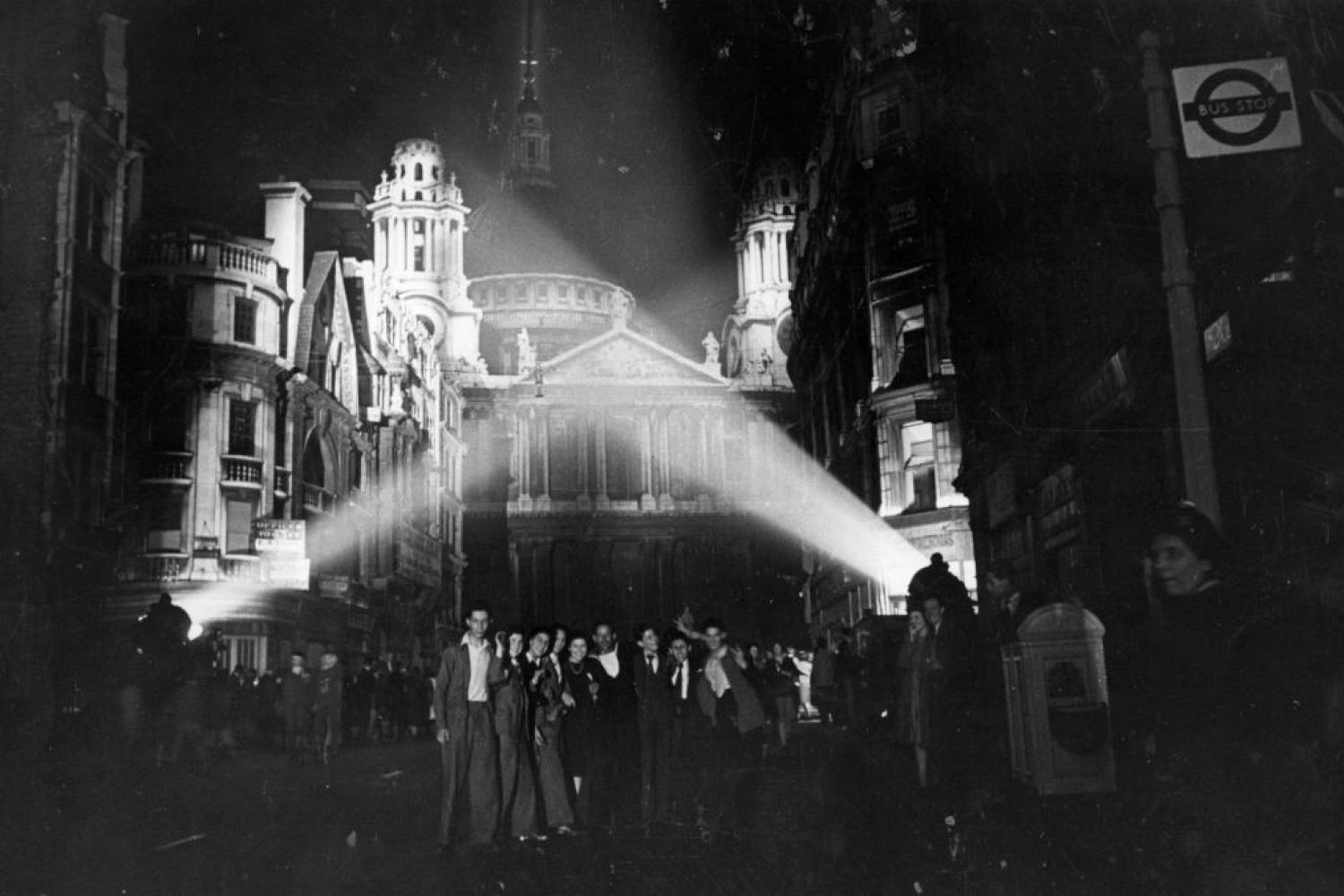 The West End of St Paul's, lit by searchlights, with a crowd of people posing for a photo in front of it.