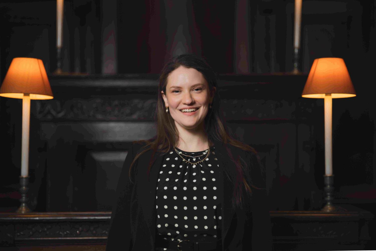 A photo of Hilary Punnet, Assistant Director of Music at St Paul's Cathedral. Hilary is a white woman with long brown hair. She is smiling at the camera, and wearing a black polka dot dress with a black blazer.