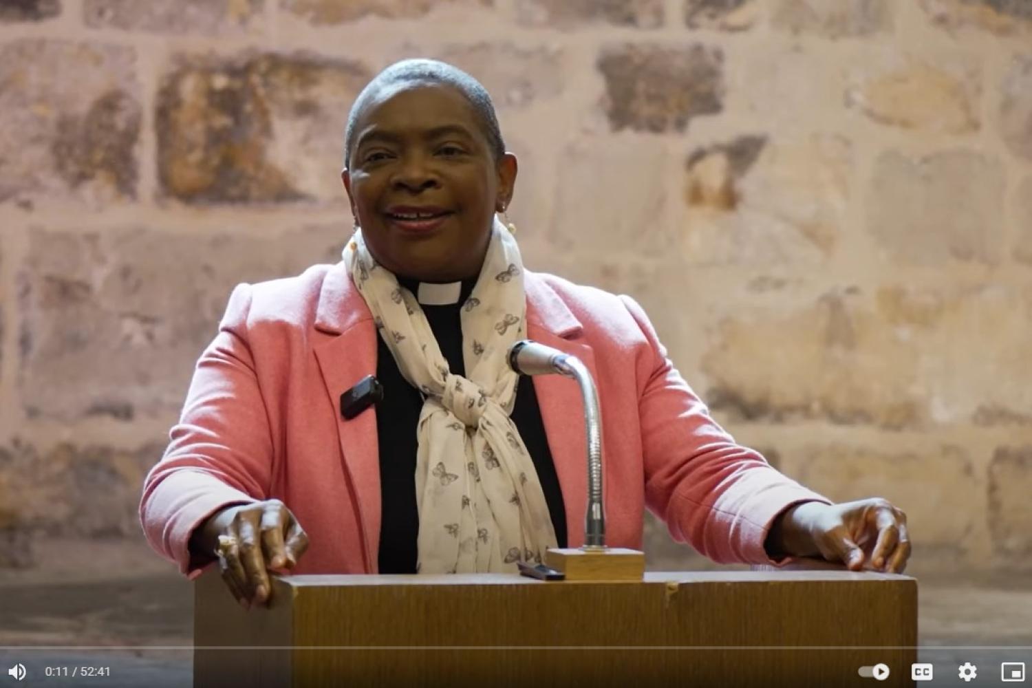 Bishop Rose is a black woman wearing a pink jacket over a black clerical shirt and a cream knotted scarf. She stands in front of a microphone and lectern in the wren suite of St Paul's Cathedral.