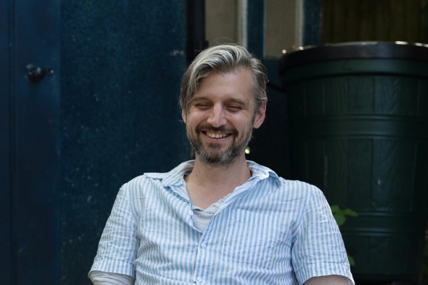 David is a white man with light brown hair and a darker beard, sitting outside in a garden chair, wearing a pale blue short sleeved shirt.