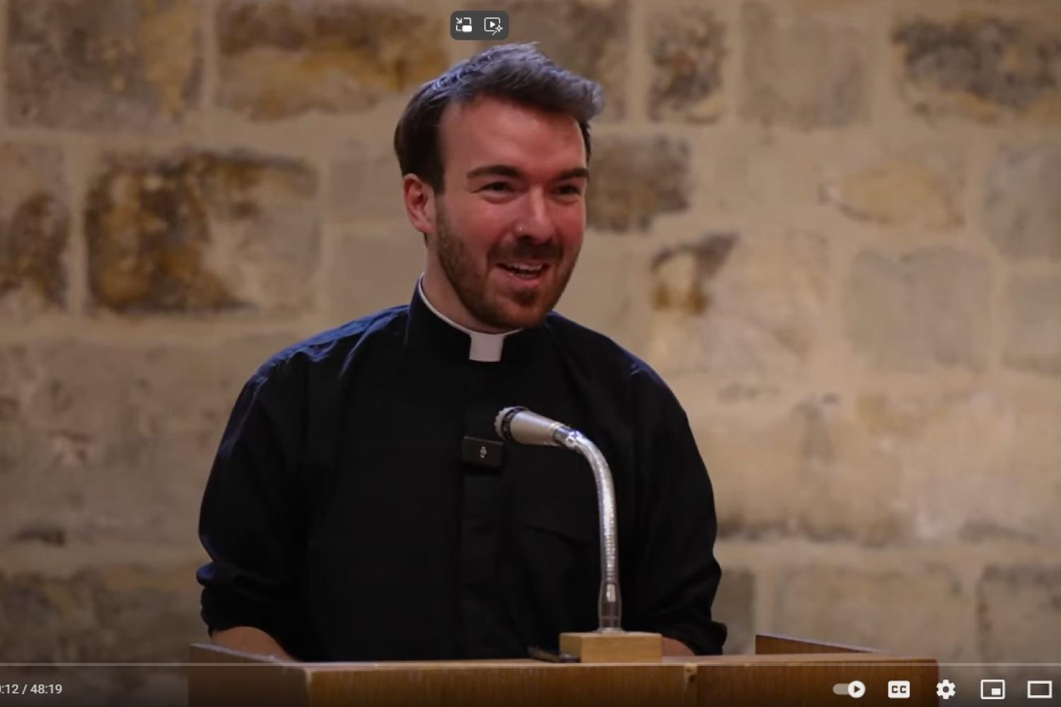 Will is a white man with short dark hair and beard, wearing a black clerical shirt and white collar, standing at a microphone in the Wren Suite at St Paul's Cathedral