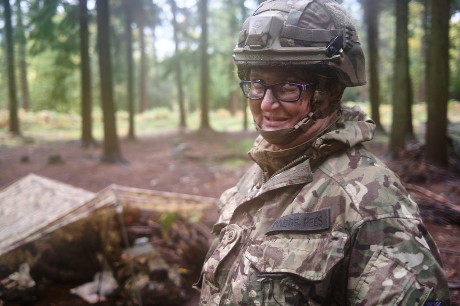 Adele wears army fatigues and a helmet and stands in front of a shelter in woodland. She wears a badge which says Padre Rees.