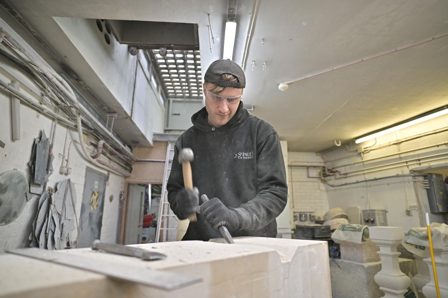 male person wearing a cap and black clothes in a workshop using a hammer and chisel to carve a stone