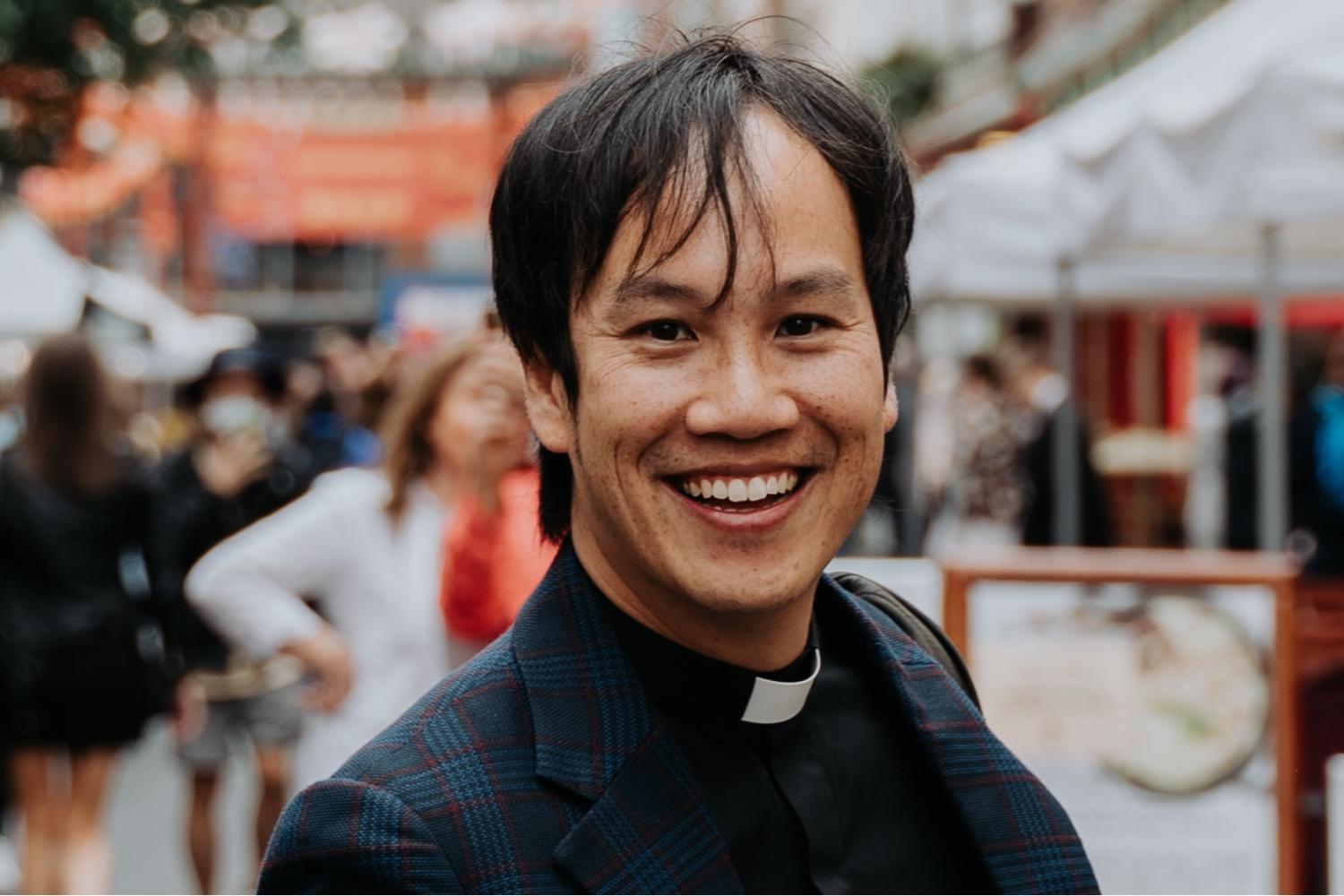 Mark Nam headshot wearing a clerical collar under a black suit and shirt, standing in a busy marketplace