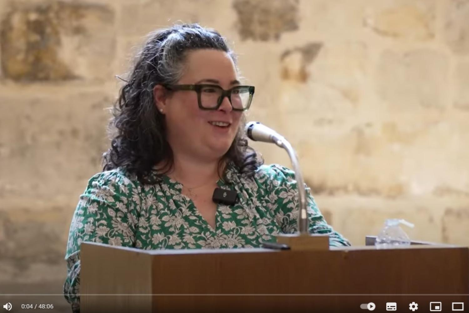 Karen wears glasses and a green and white patterned dress and stands at a lectern with microphone