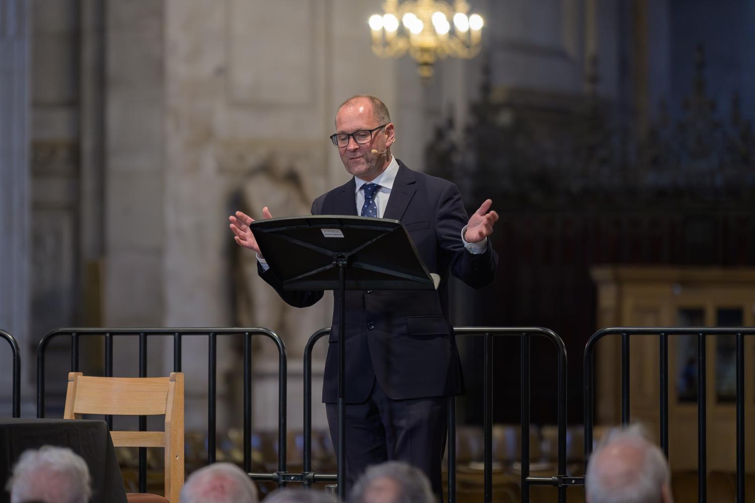 Allen stands at a lectern on a stage in St Paul's Cathedral, arms out in front of him as he speaks to an audience