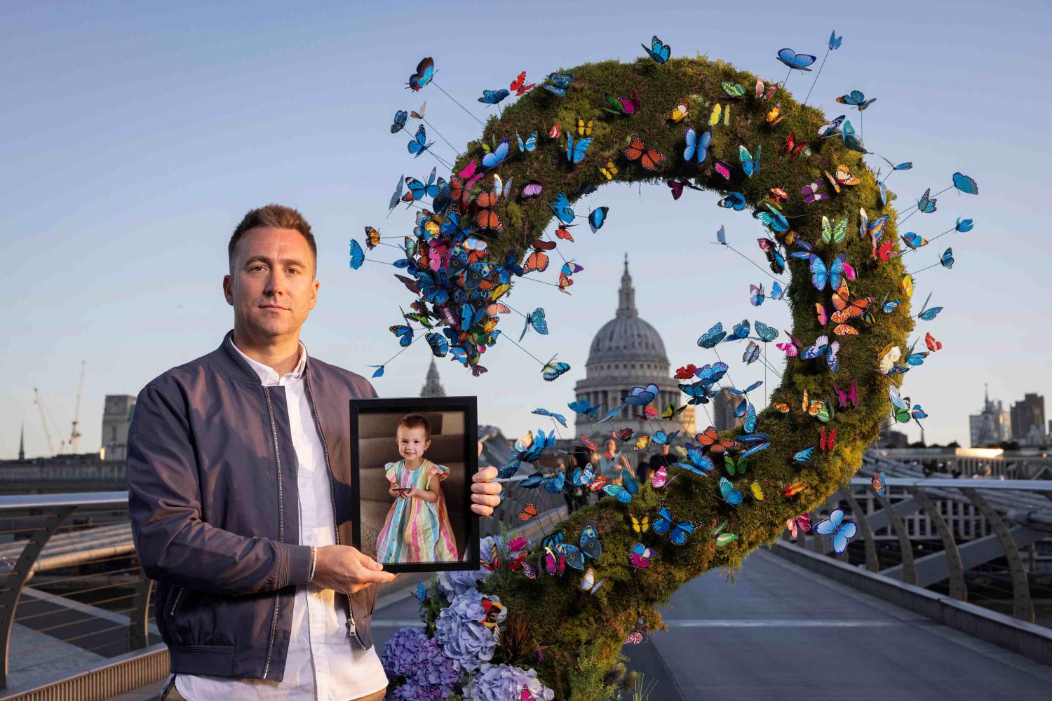 A man stands on Millennium Bridge in London, dressed in a white shirt and blue jacket, with short blond hair. He is holding a photo of his late daughter, who passed away from cancer. He is standing beside an art work for Great Ormond Street Hospital Charity, made of butterflies, framing the dome of St Paul's Cathedral.