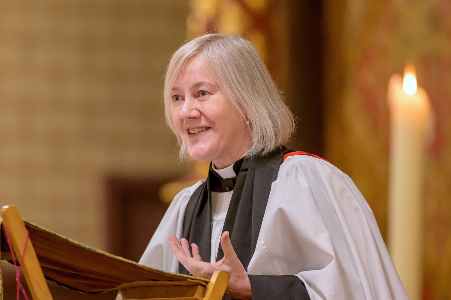 Ellen wears a clerical collar and robes while standing at a lectern, her hand outstretched as she talks