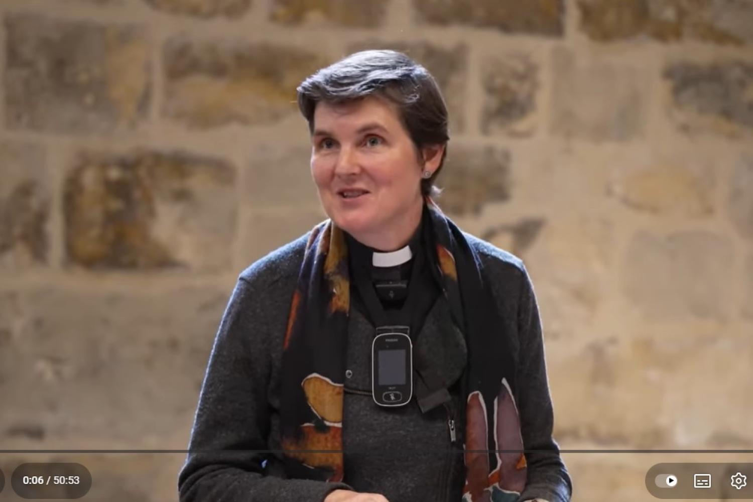 A still from the film in which Morwenna stands in the Wren Suite speaking to an audience