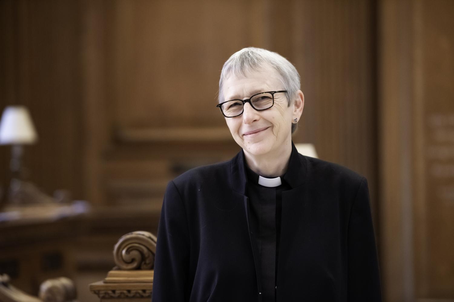 Alison wears a black jacket and shirt with a clerical collar and is photographed inside a church