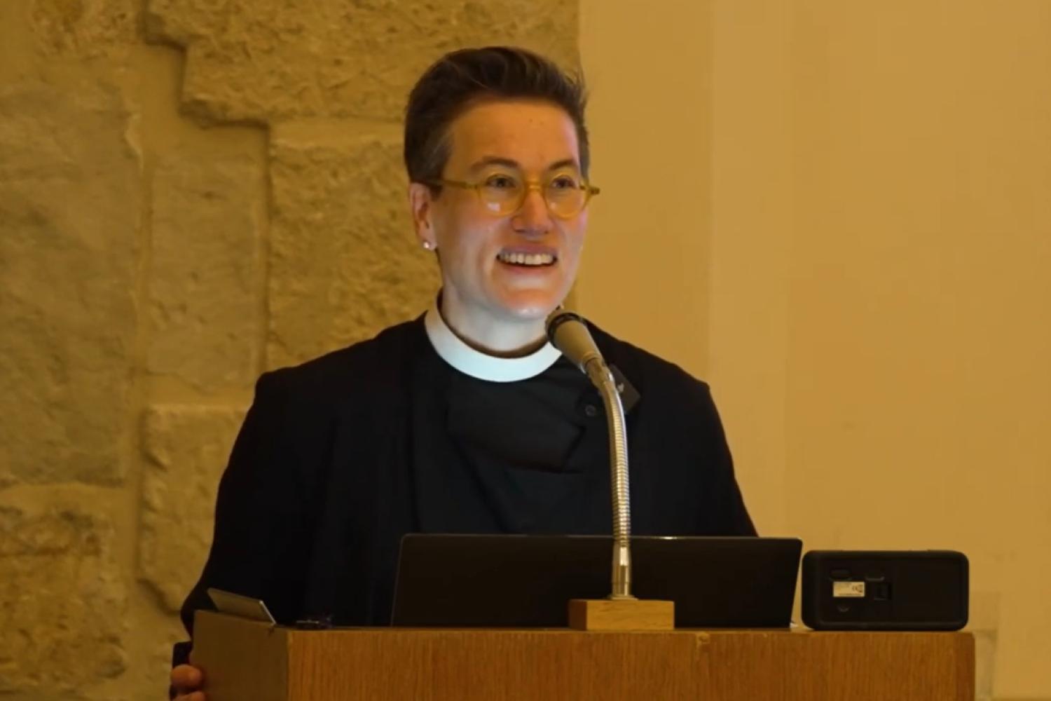 Ayla wears a clerical collar and black shirt and stands at a microphone and lectern