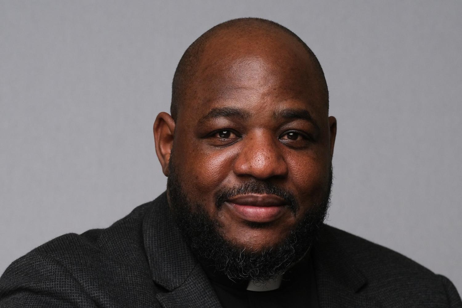 Carlton wears a clerical collar with a black shirt and jacket