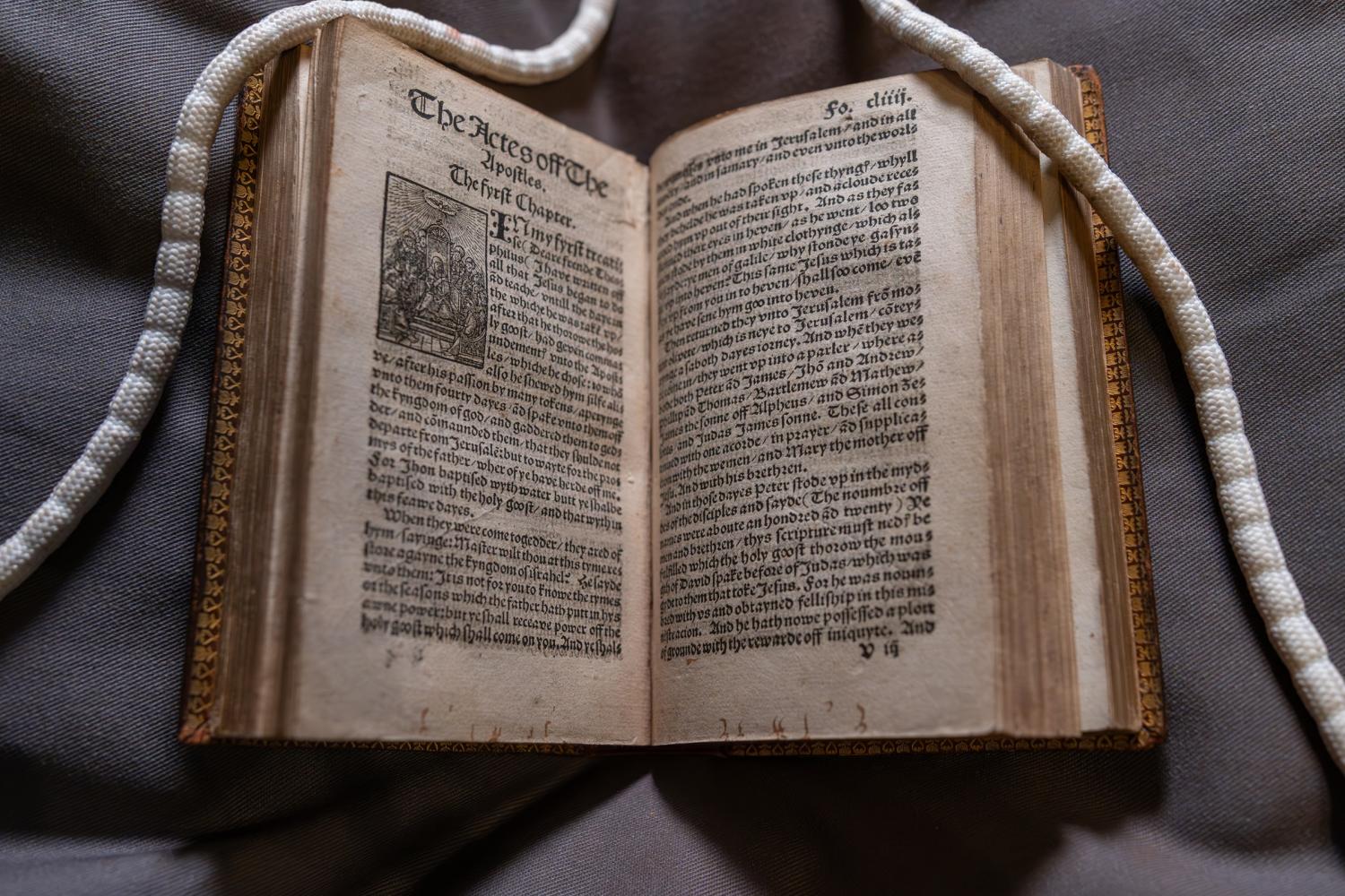 Tyndale Bible open at "The Acts of the Apostles". Long white "snake" weights are visible holding the top right and left corners of paper open. 