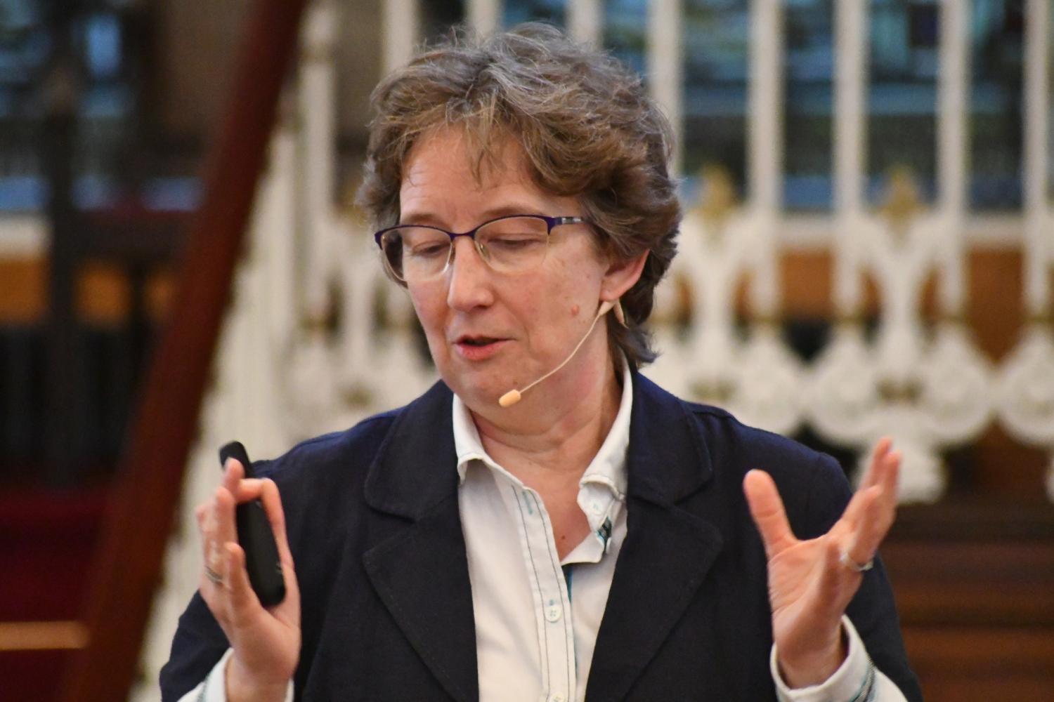 Helen has short brown hair and glasses, and stands with hands raised and wearing an ear microphone as she talks.
