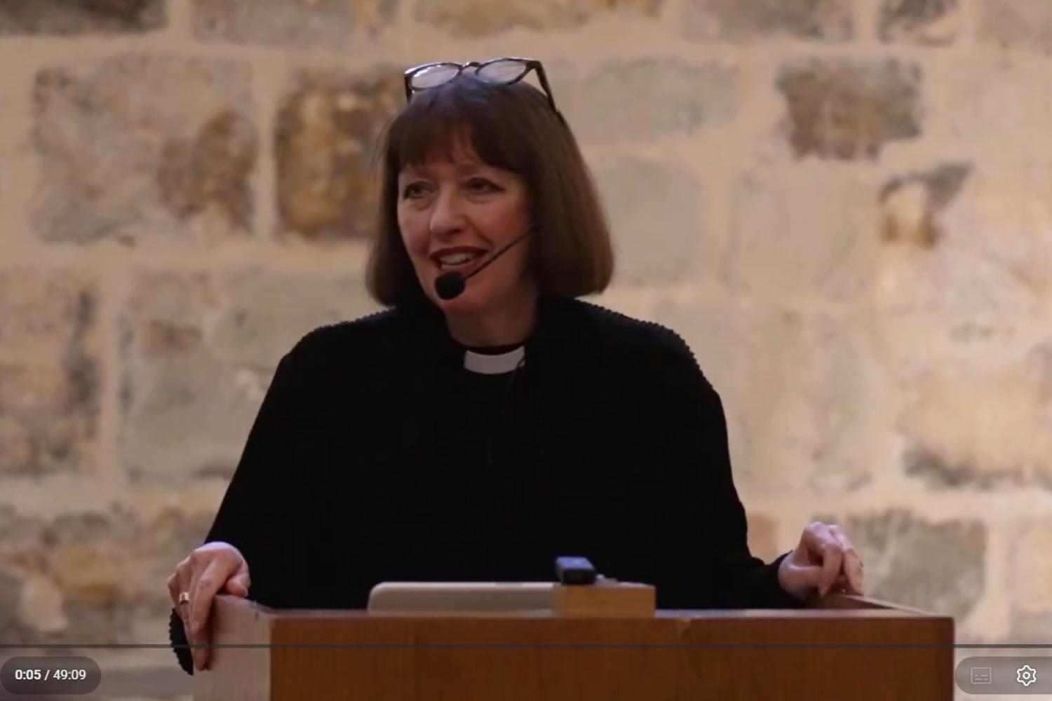 Carys Walsh, wearing a black shirt and clerical collar, with an ear microphone and glasses on her head, stands at a lectern in the Wren Suite at St Paul's Cathedral.
