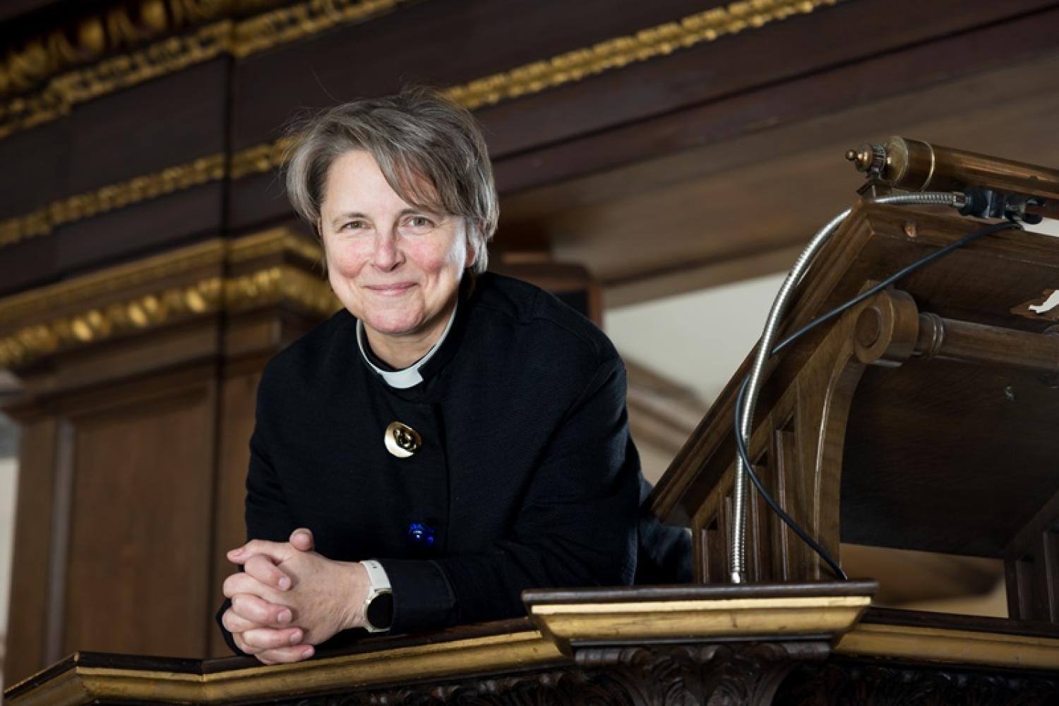 Lucy wears a clerical collar and black shirt, has short brown hair, and leans against a wooden pulpit in a church