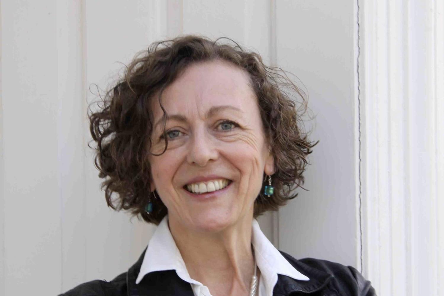 Maggi has short wavy brown hair and wears a white shirt under a black jacket