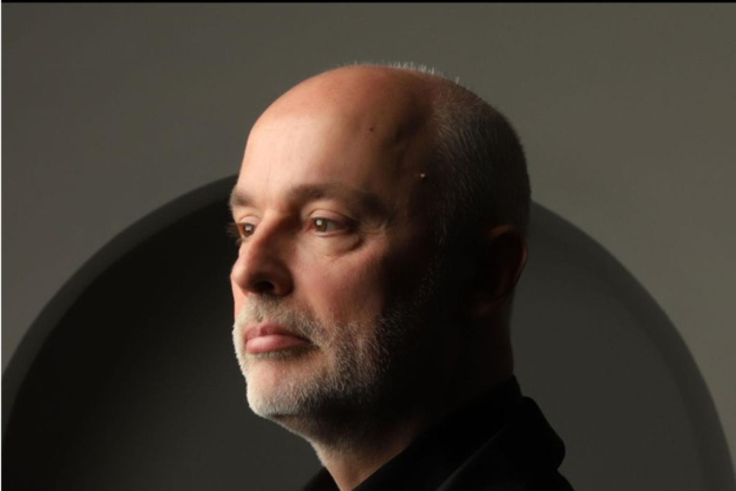 Patrick has short grey hair and a grey beard, wears a black suit and shirt and faces to the left