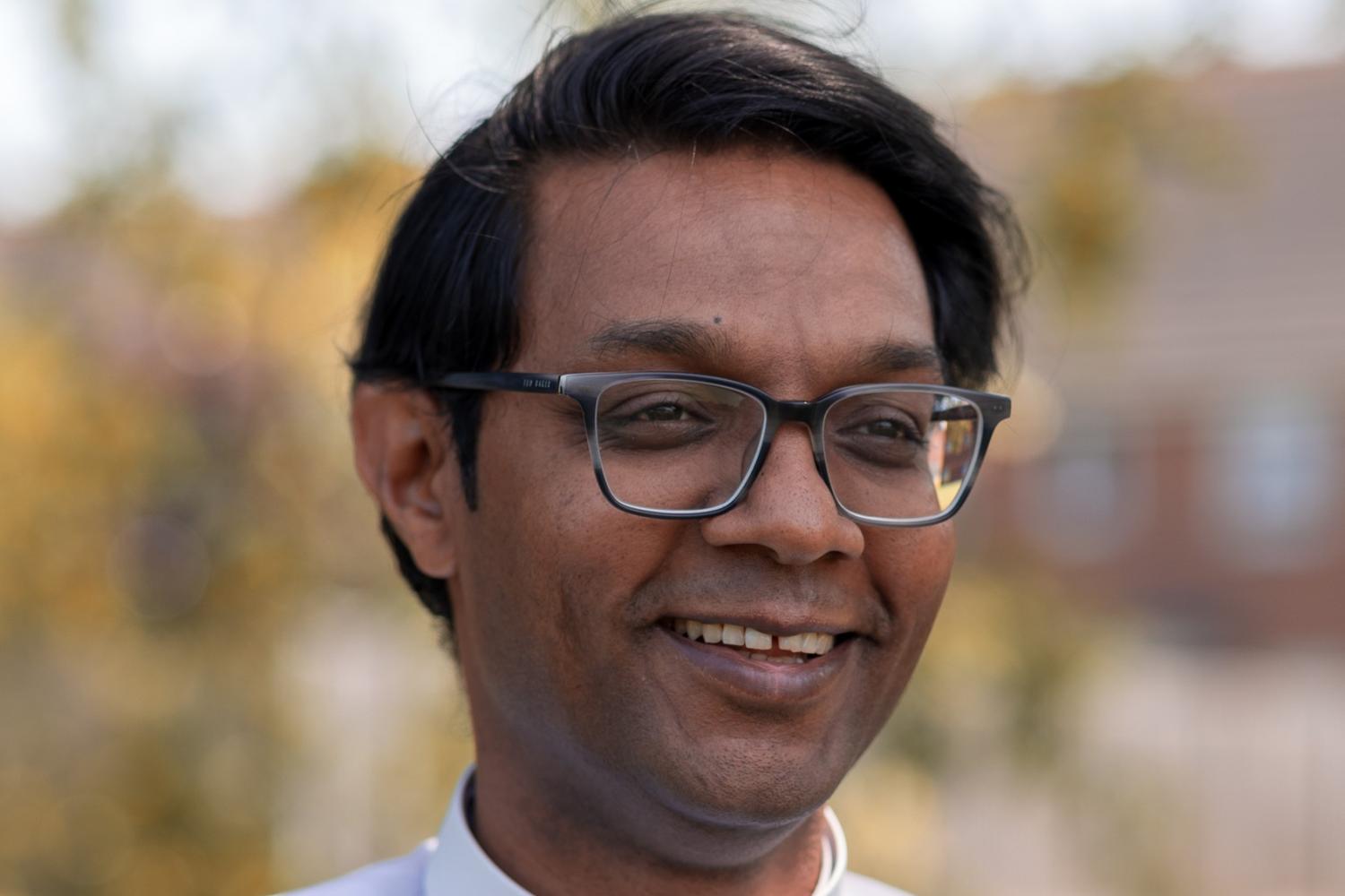 Peniel wears a clerical collar with a pale blue shirt and stands in the shade of a tree