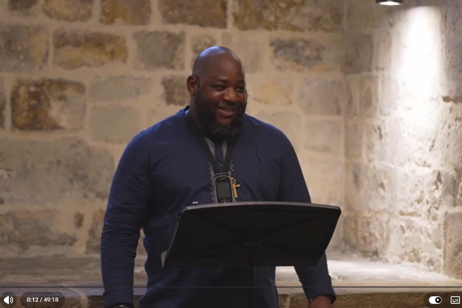 Carlton wears a dark blue top and stands before a lectern in the Wren Suite in St Paul's Cathedral