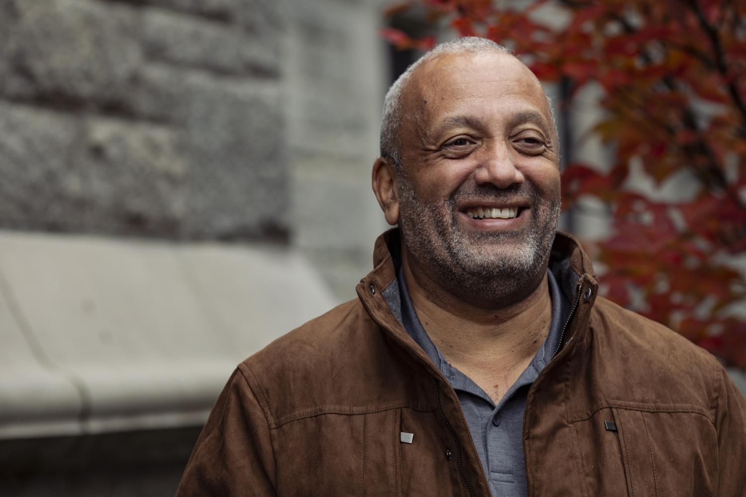 John has short grey hair, wears a brown jacket over a grey shirt, and stands in front of a church building