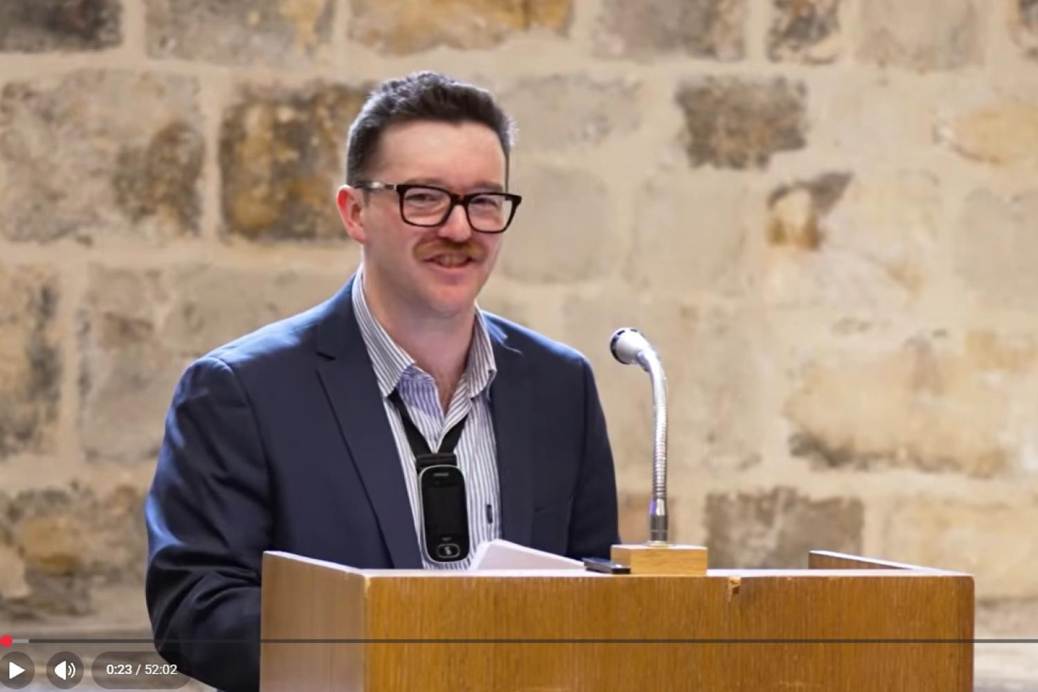 Michael has short auburn hair and a moustache, and wears black framed glasses. Wearing a navy suit and striped blue shirt he stands in front of the lectern in the Wren Suite of St Paul's Cathedral