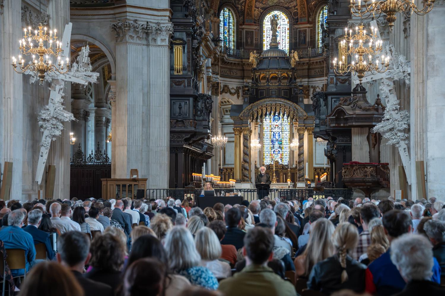 A large audience in the nave of the cathedral with the quire and high altar in the background and a stage visible with two speakers