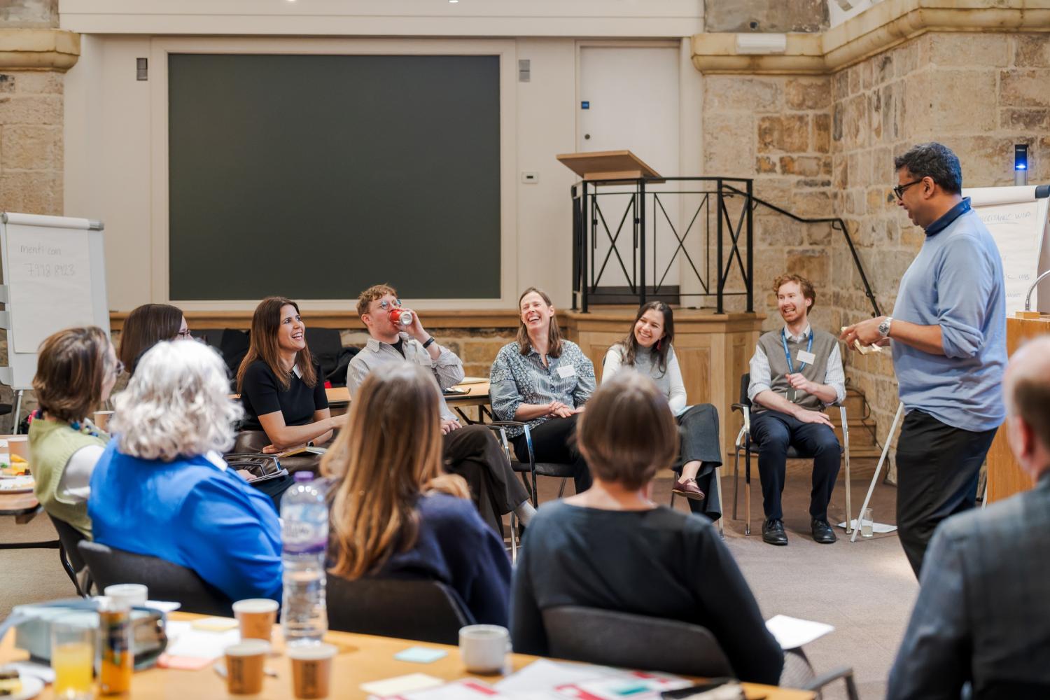 A group of seated people laugh as a facilitator stands in front of them