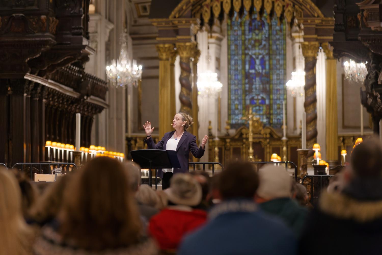 Paula Gooder stands on a stage with hands raised as she talks with a large audience in front and the quire and high altar in the back