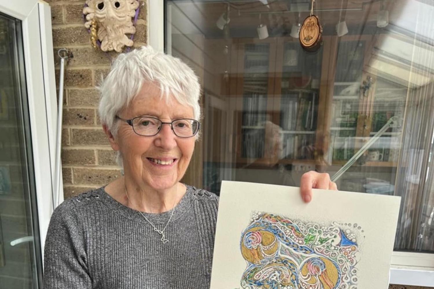 A woman with short white hair and glasses standing in a conservatory holding a finished piece of art decorated with gold leaf and pastel colours