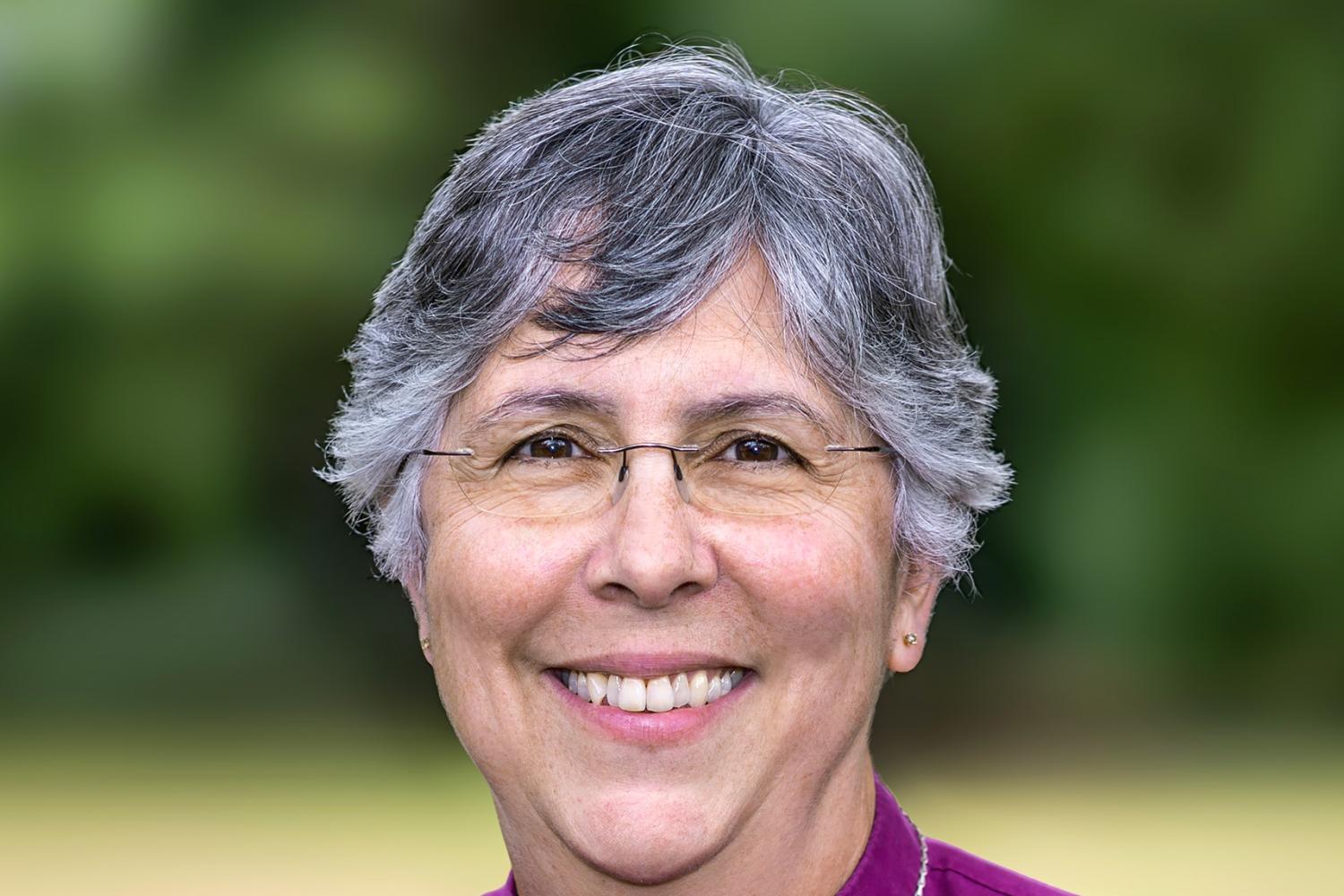a woman with short grey hair and glasses wears a purple shirt and clerical collar with a large cross on a chain