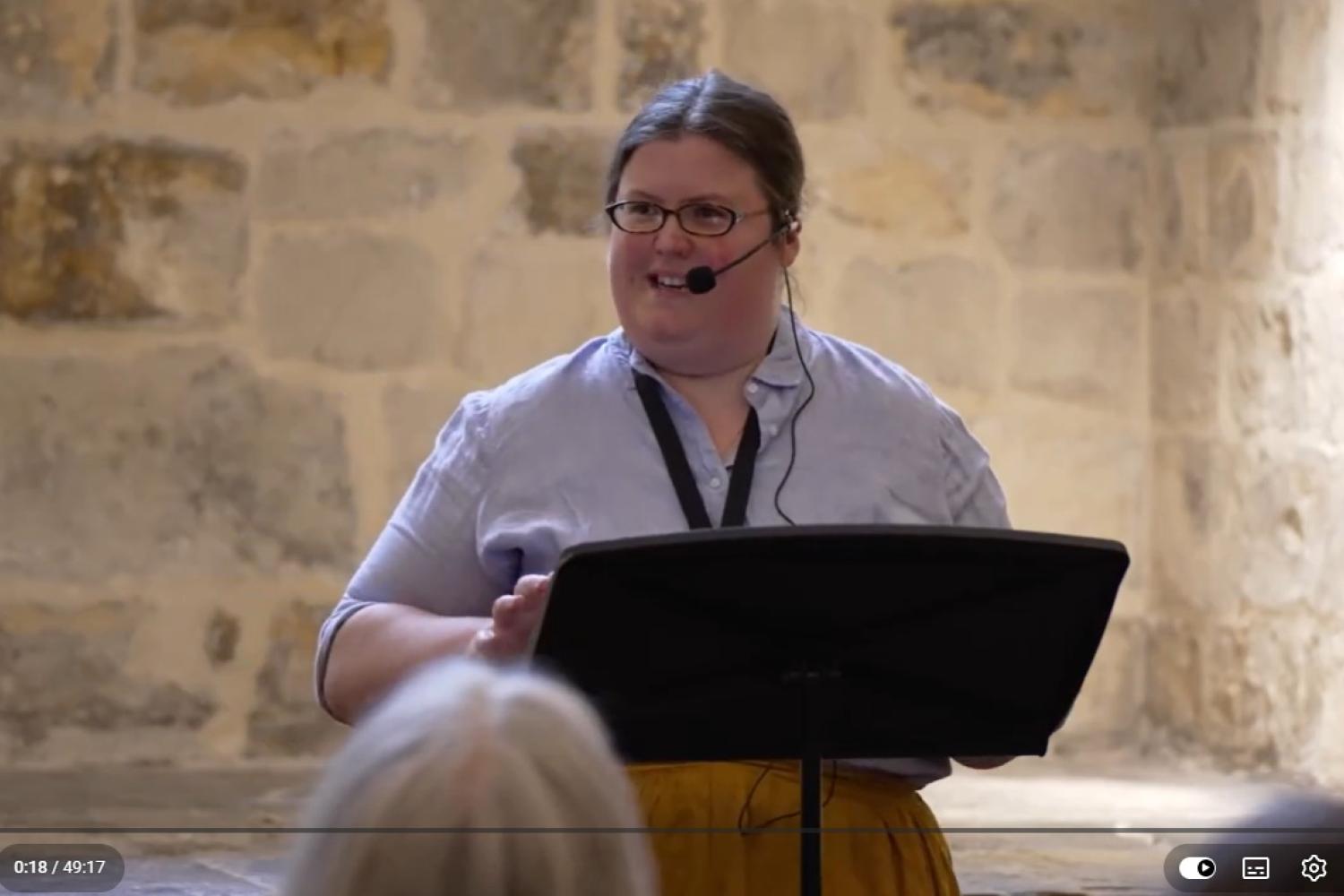 Charlotte has long hair in a pony tail and glasses, and wears a pale blue shirt and stands at a lectern in the Wren Suite in St Paul'[s Cathedral