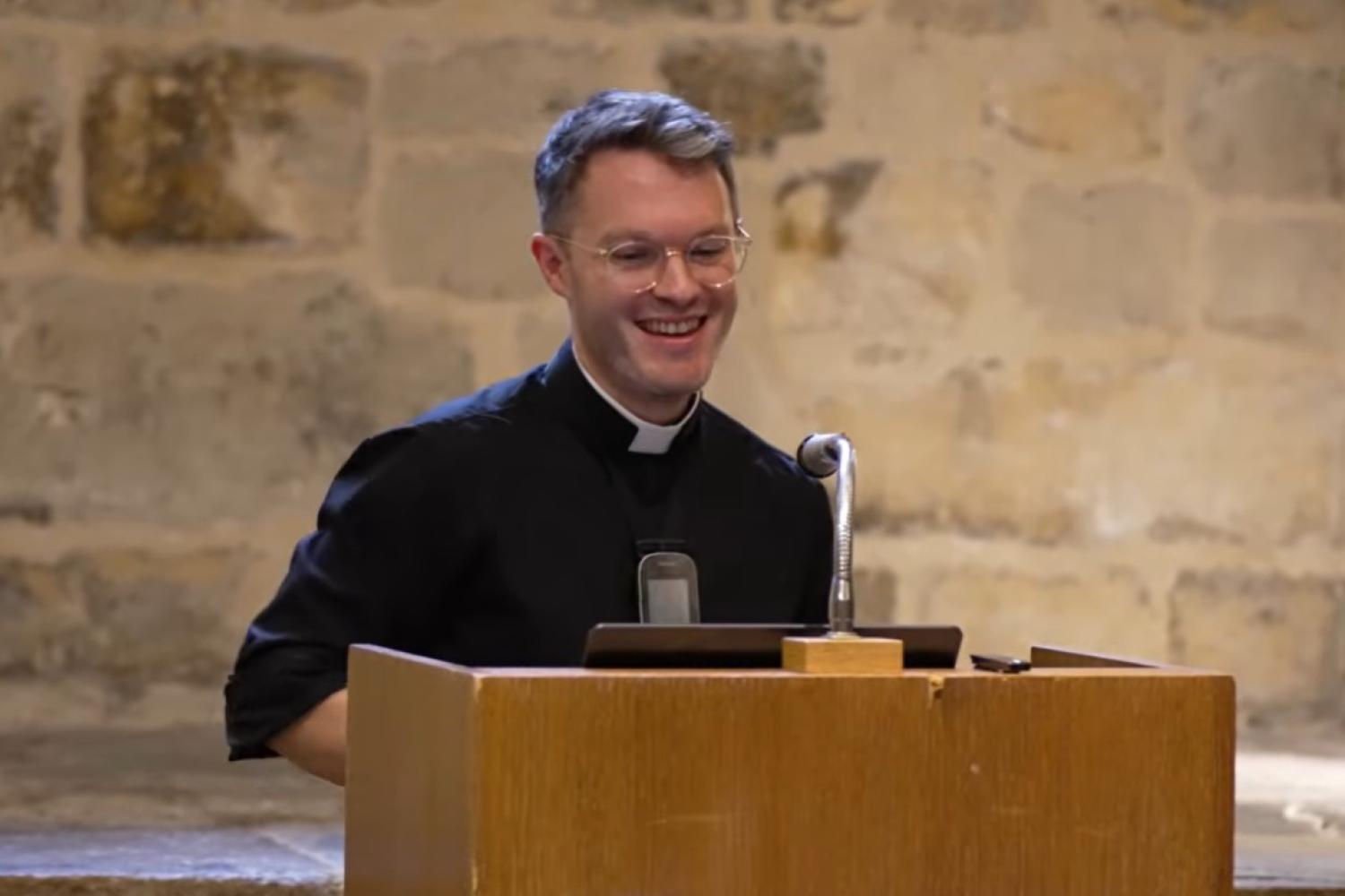 Steffan has short brown hair and glasses, wears a clerical collar and a black shirt, and stands at a lectern in the Wren Suite in St Paul's Cathedral