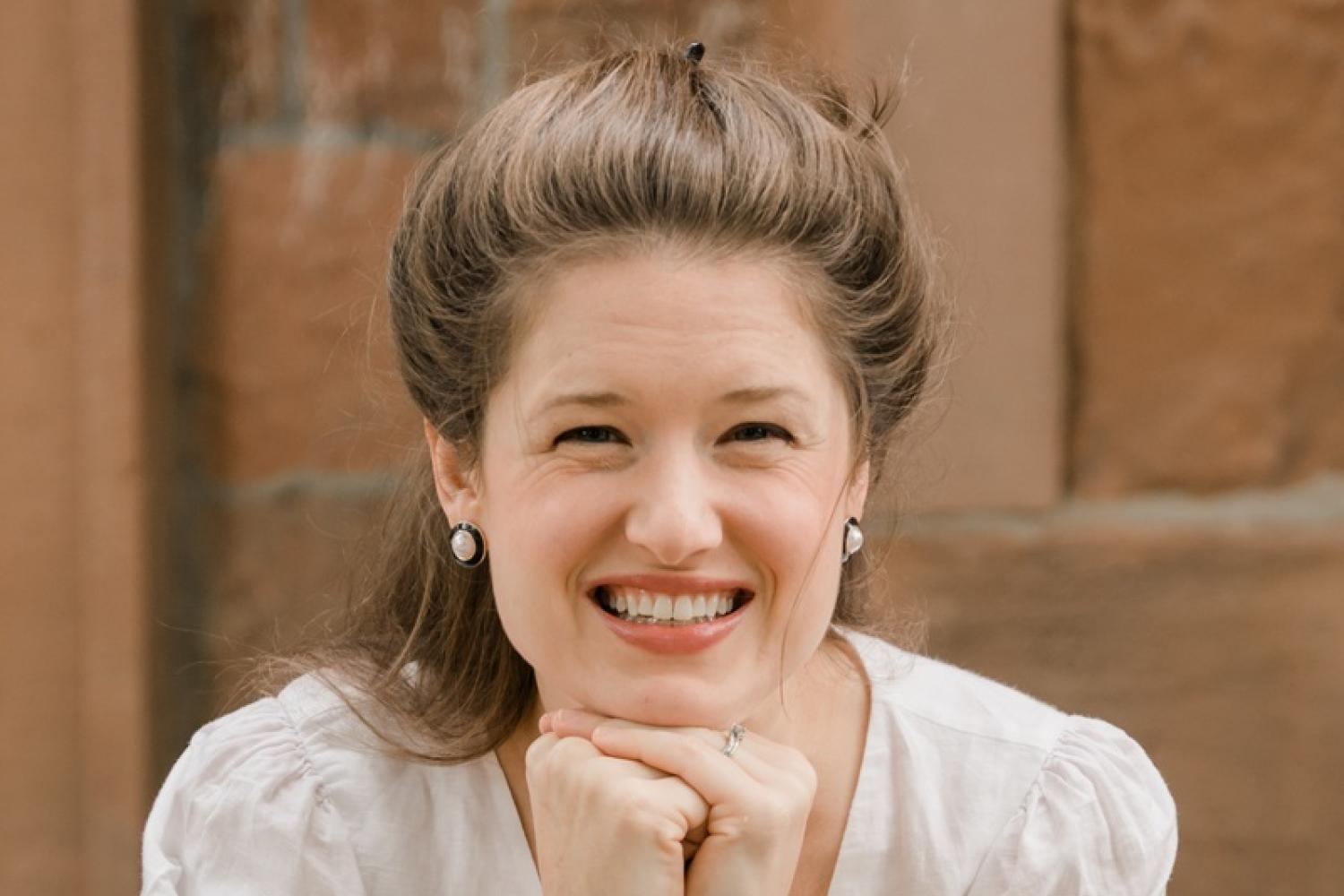 A woman with long light brown hair pulled into a bun sits on steps outside an old brick building, her chin resting on her hands
