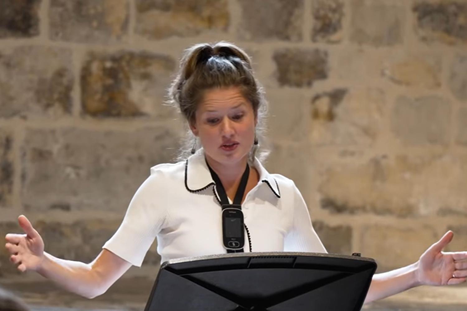 A woman with long hair in a high pony tail wearing a white blouse stands at a lectern with arms outstretched as she talks in a room with old exposed stone walls