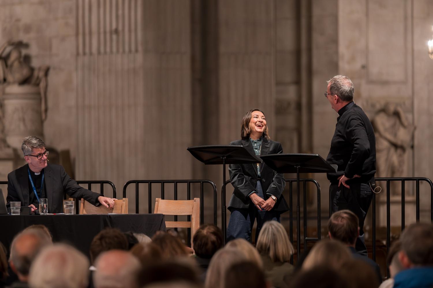 A woman and man stand on a stage, and she is laughing as they talk. Another man sits at a table alongside them, smiling. The portland pillars of the cathedral can be seen behind the stage.