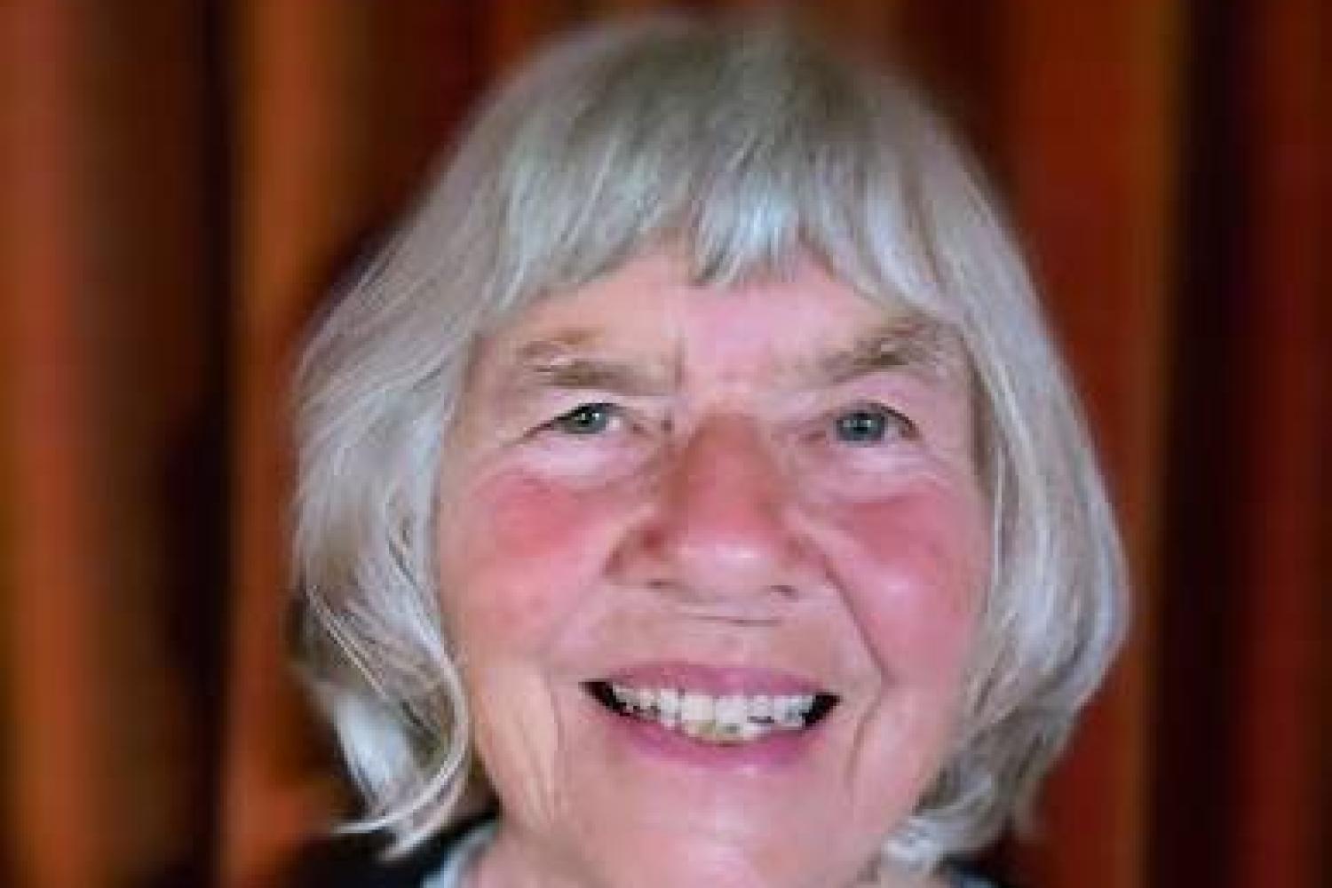 Margaret has short grey hair, wears a flowery top under a black jacket, an smiles at the camera