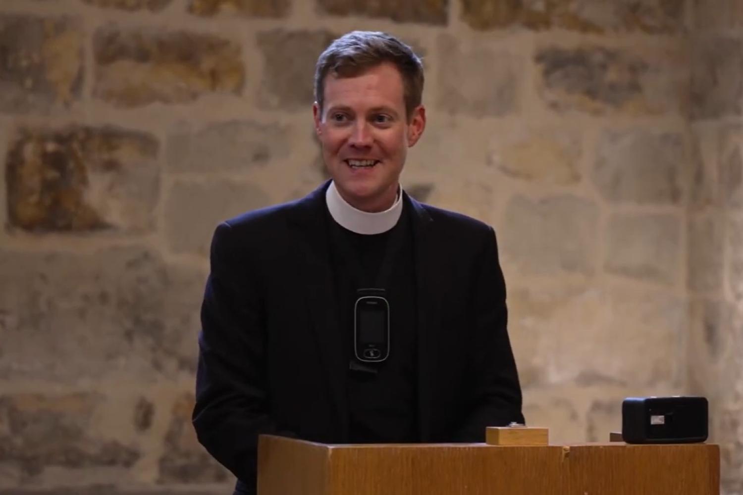 Simon wears a clerical collar under a black suit and has short auburn hair. He stands at a lectern in the Wren Suite in the crypt of St Paul's Cathedral