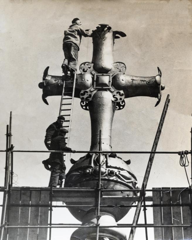 A black and white photograph of men working on the ball and cross with ladders and scaffolding