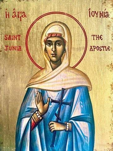 A picture of the icon of St Junia. She is depicted wearing a blue robe and a white headscarf trimmed with red, and she is dark-skinned with dark brown eyes. She has one hand raised with her palm facing the viewer and holds a dark blue cross in the other hand. The cross has a smaller slanted crossbeam below the main crossbeam. She has detailed gold and white cuffs at the end of red sleeves, or alternatively may be wearing decorative bangles. She is painted on a gold leaf background with a red double-ringed h